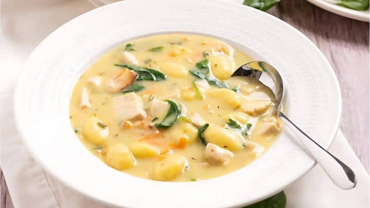 A bowl of creamy soup with gnocchi, chicken pieces, spinach, and carrots, served with a spoon on a white plate.
