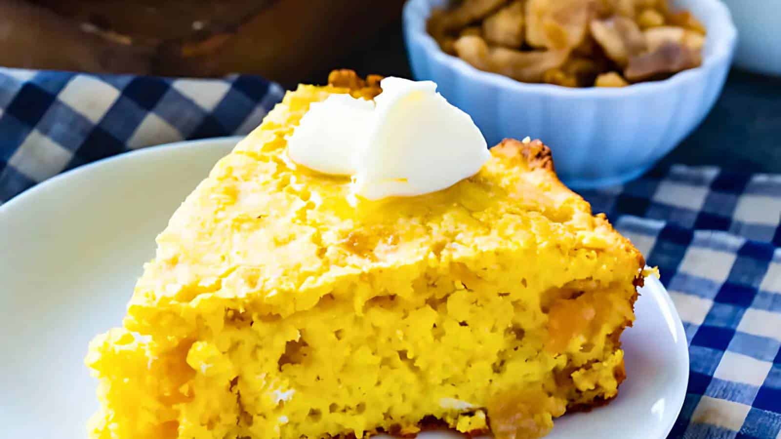 A slice of cornbread on a white plate with a dollop of butter on top. A bowl with nuts is in the background, and a blue and white checkered cloth is partially visible.