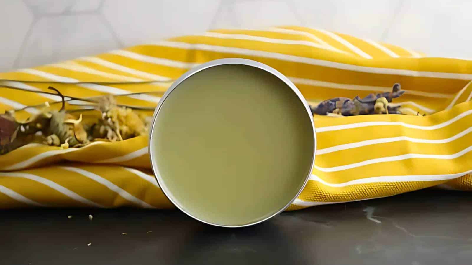 A round tin of green balm is placed on a black surface in front of a yellow and white striped cloth with dried flowers around it.