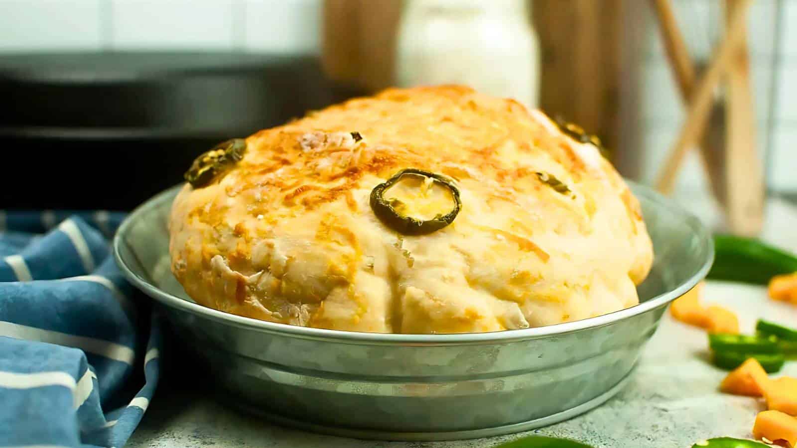 A round loaf of bread topped with melted cheese and jalapeño slices in a metal baking dish on a countertop. A blue striped cloth and some chopped vegetables are nearby.
