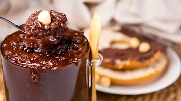 A spoonful of chocolate Nutella spread is being lifted from a jar. A hazelnut is on top. In the background, bread with chocolate spread is visible on a plate.