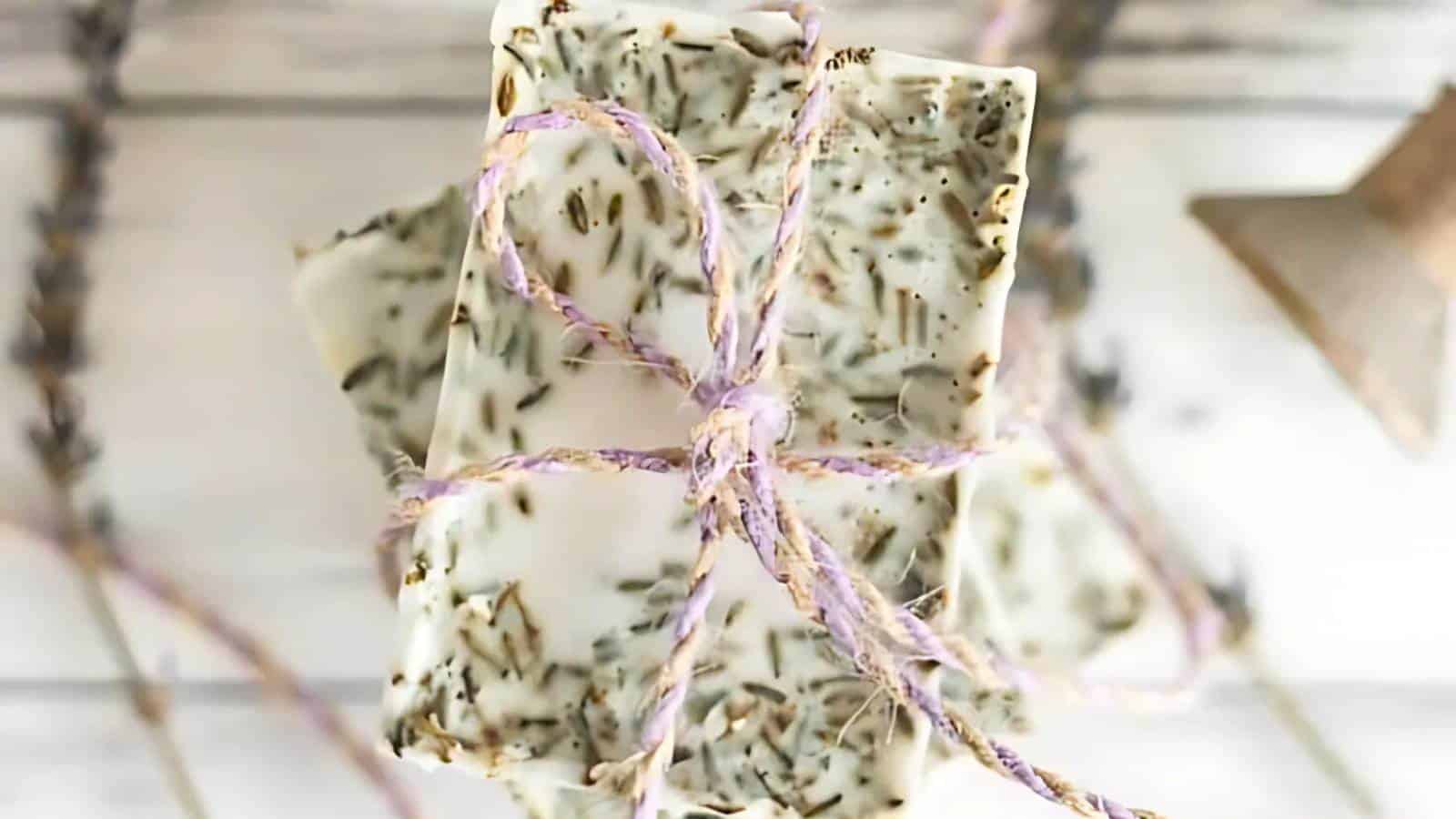 Two bars of handmade soap with embedded herbs, tied together with a purple and white twine, placed on a white wooden surface.