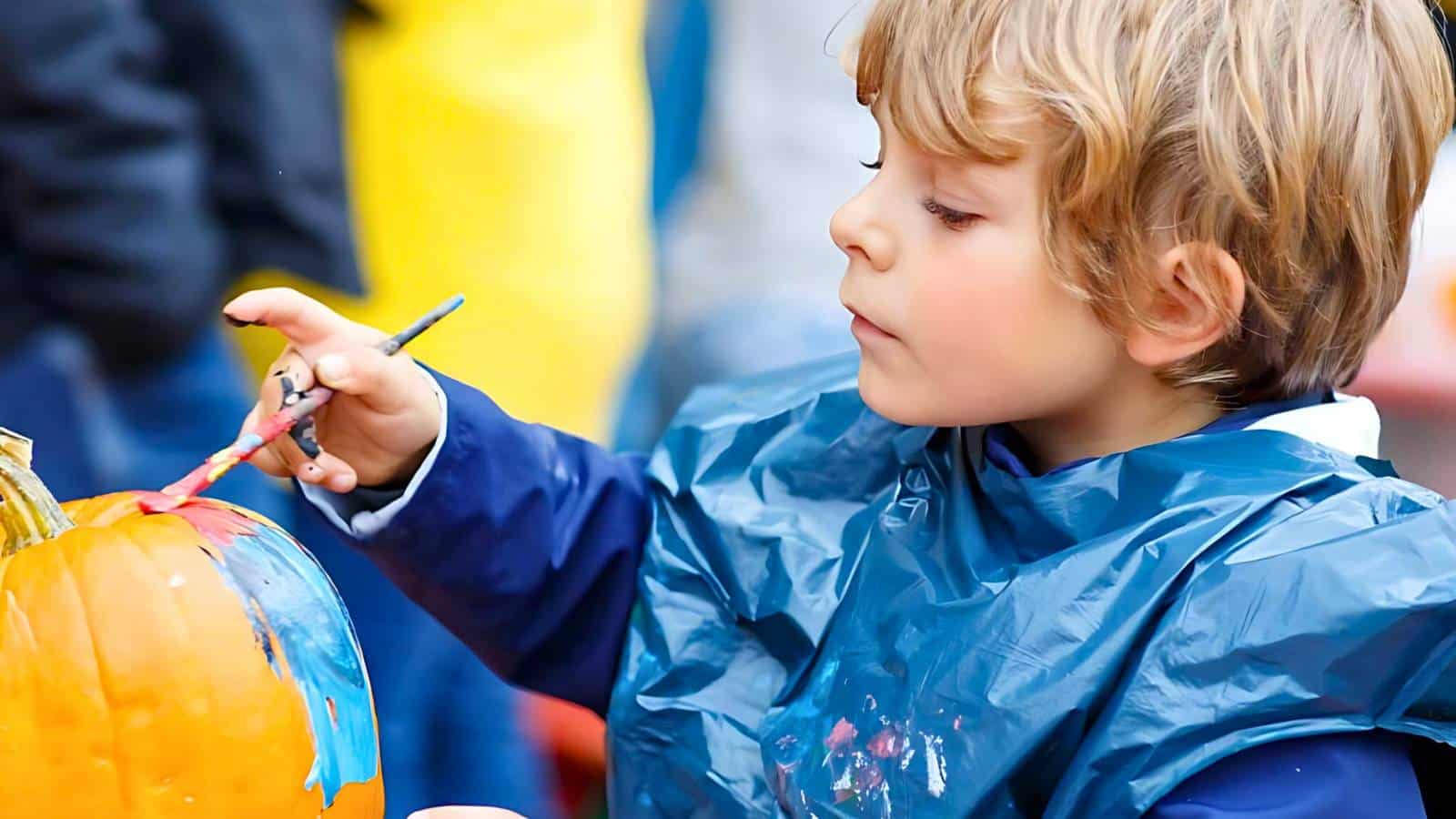 A young child in a blue smock paints a pumpkin with a small brush, applying red and blue paint.