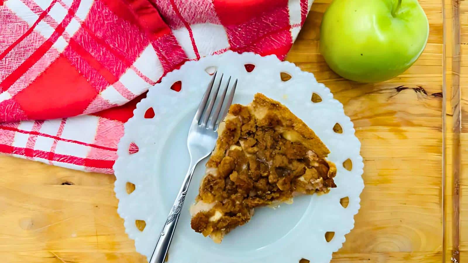 A slice of apple pie with a crumb topping on a white plate, a silver fork beside it, with a red checkered cloth and a green apple on a wooden table.
