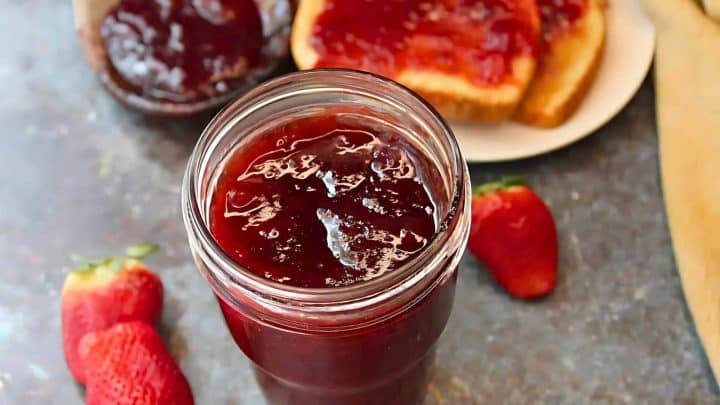 A jar of strawberry jam is open on a table with fresh strawberries nearby. Two slices of bread with jam are on a plate in the background.
