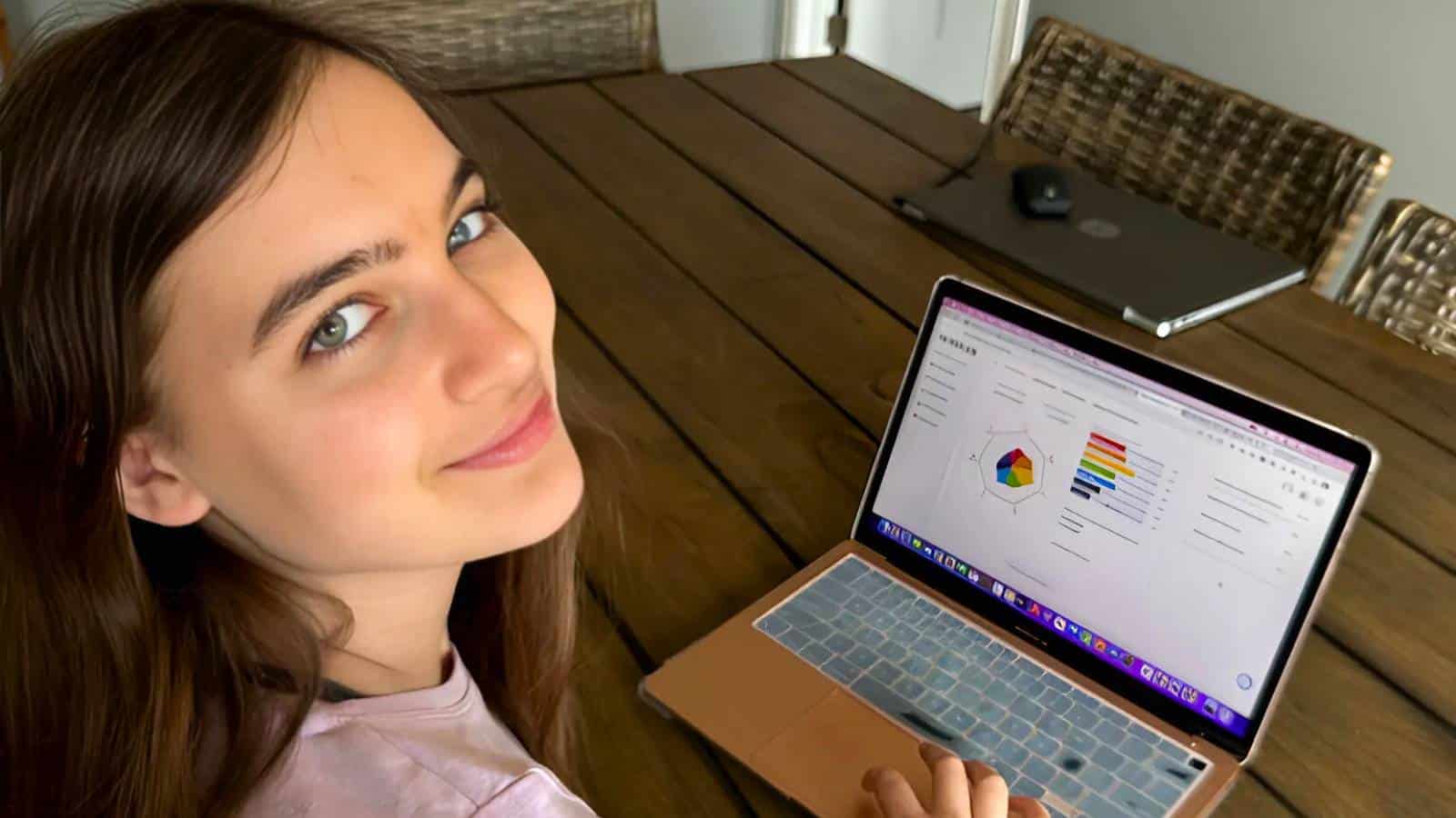 A person sitting at a wooden table looks at the camera while using a laptop displaying charts and graphs.