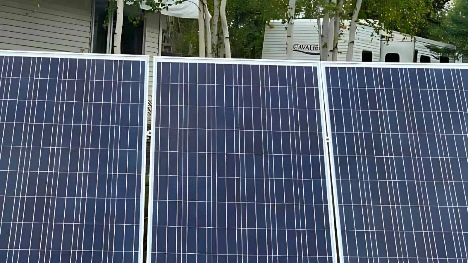 Close-up view of three solar panels angled towards the sun, with trees and a white camper in the background.