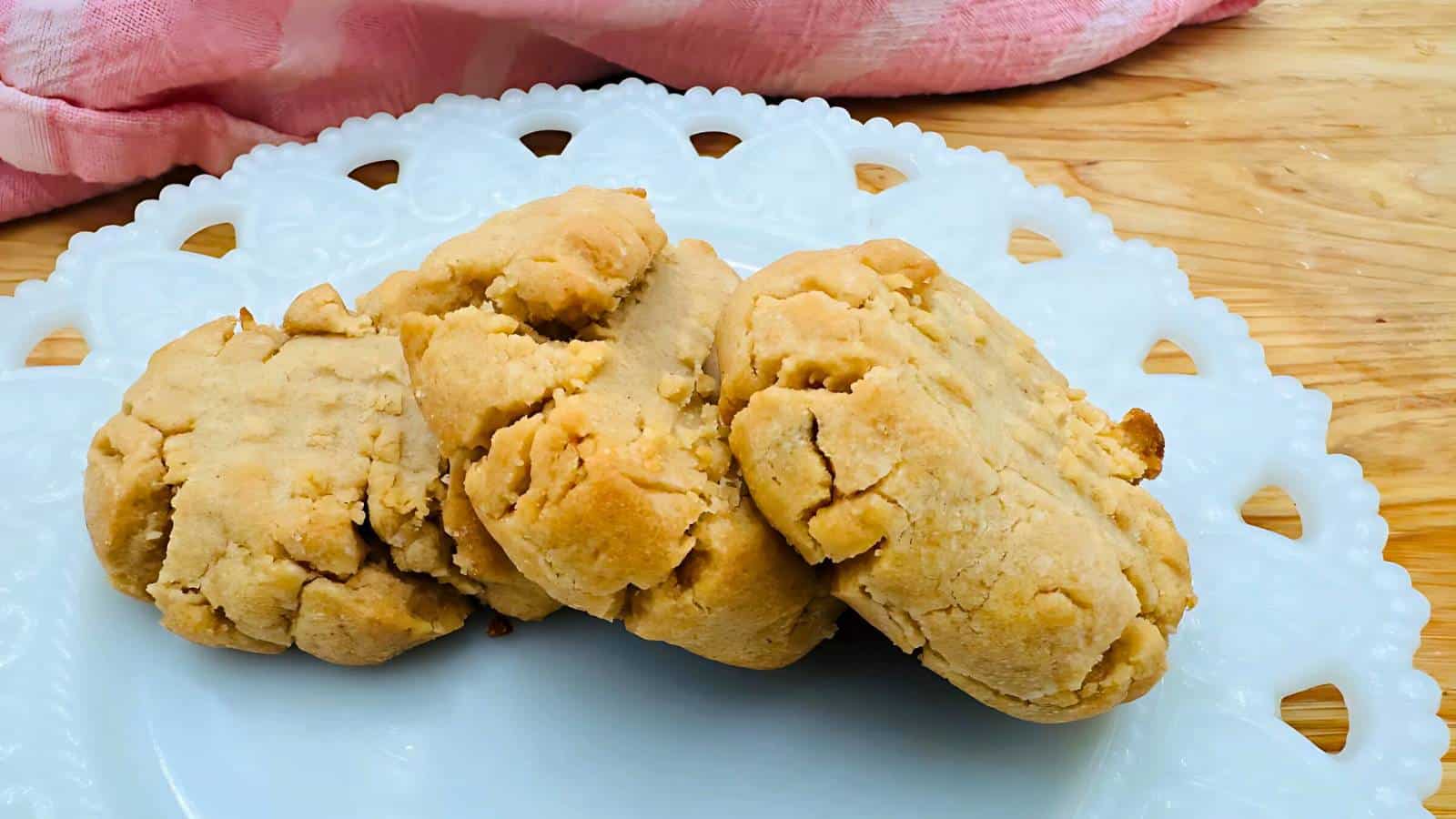 Four peanut butter cookies on a white decorative plate with a pink cloth in the background.