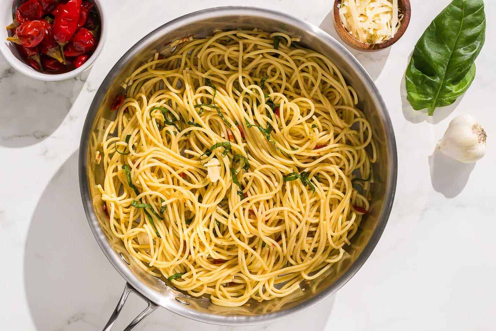 A pan of plain spaghetti with herbs, surrounded by a bowl of roasted red peppers, a small bowl of grated cheese, a garlic bulb, and a basil leaf on a white surface.