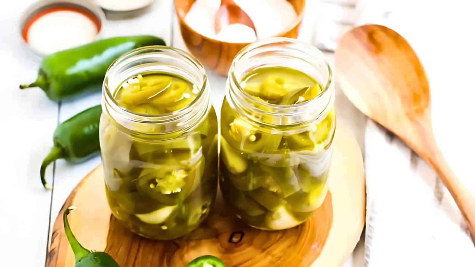 Two glass jars filled with sliced pickled jalapeños on a wooden board, surrounded by whole jalapeños, a wooden spoon, and some small bowls.