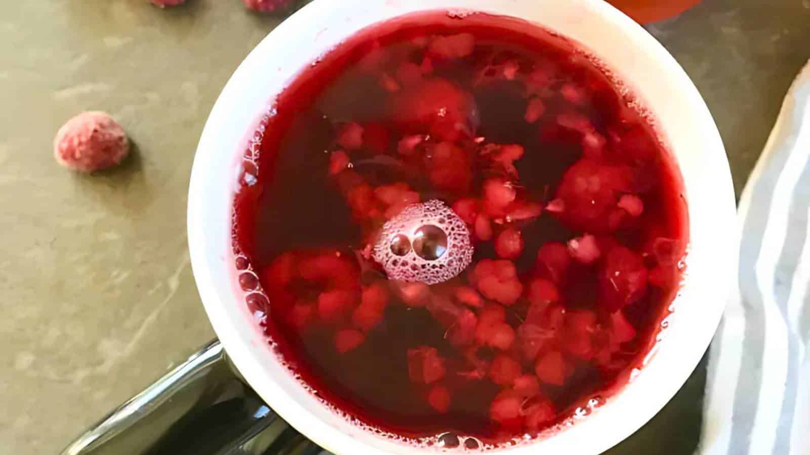 Cup filled with red liquid containing fruit pieces, viewed from above. A frozen raspberry is on the table beside the cup.