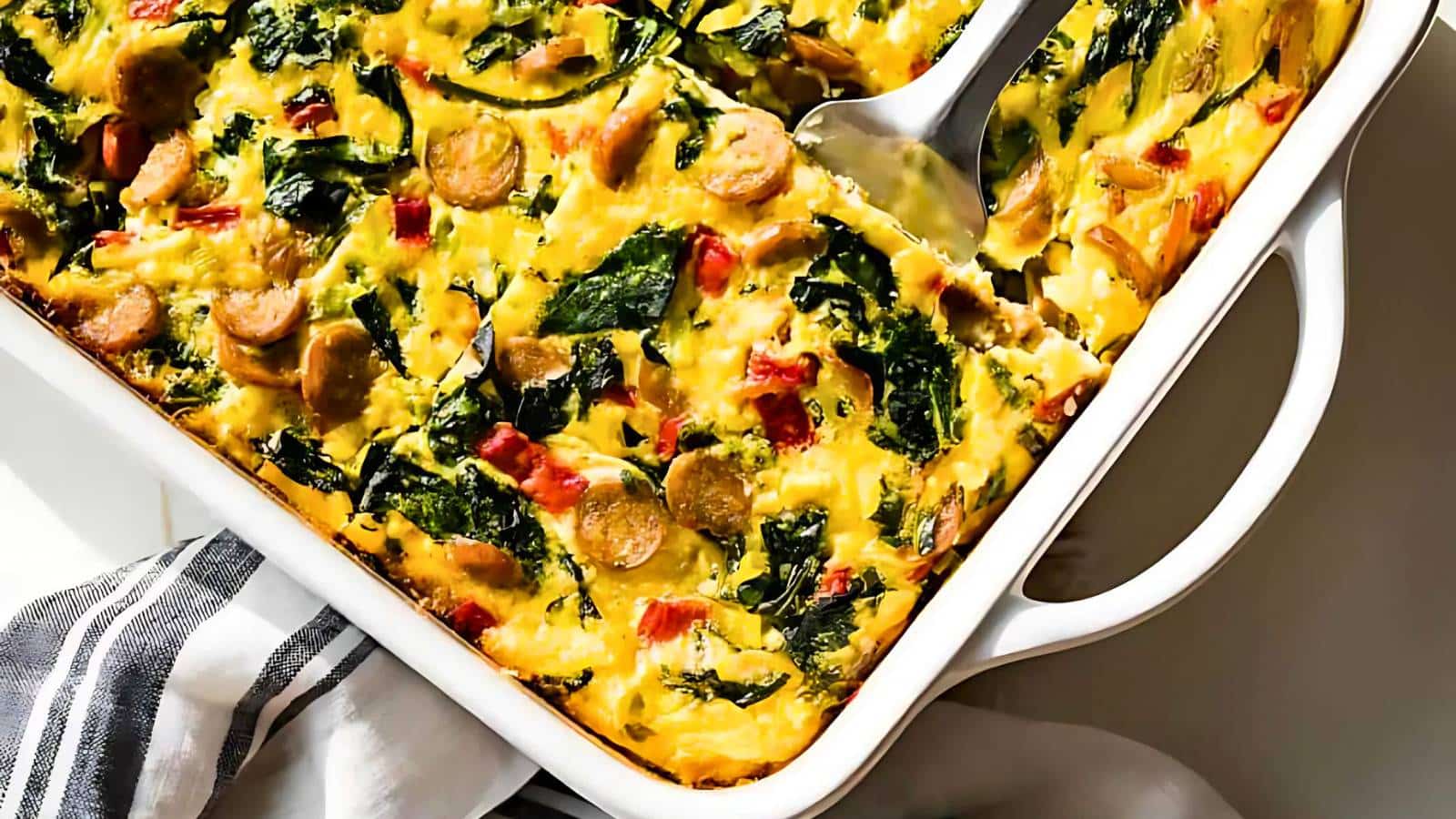 A baked casserole with sausage slices, spinach, and diced red peppers, served in a white dish with a checkered towel beneath. A serving spatula is lifting a portion.