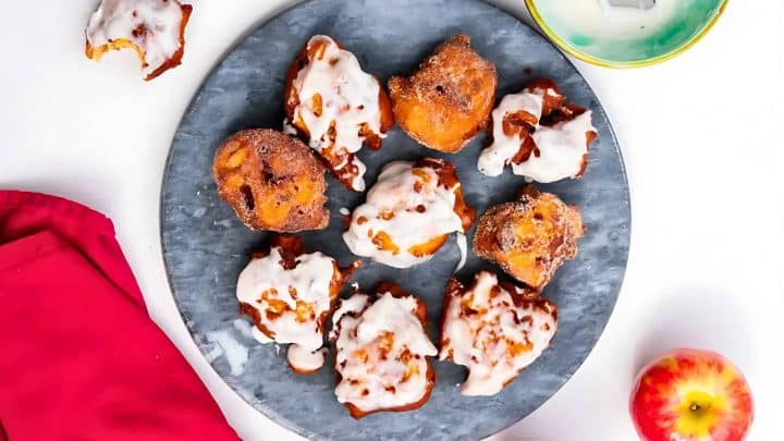 A plate of apple fritters covered in white glaze is displayed alongside a red napkin, a whole apple, and a partially visible yellow bowl.