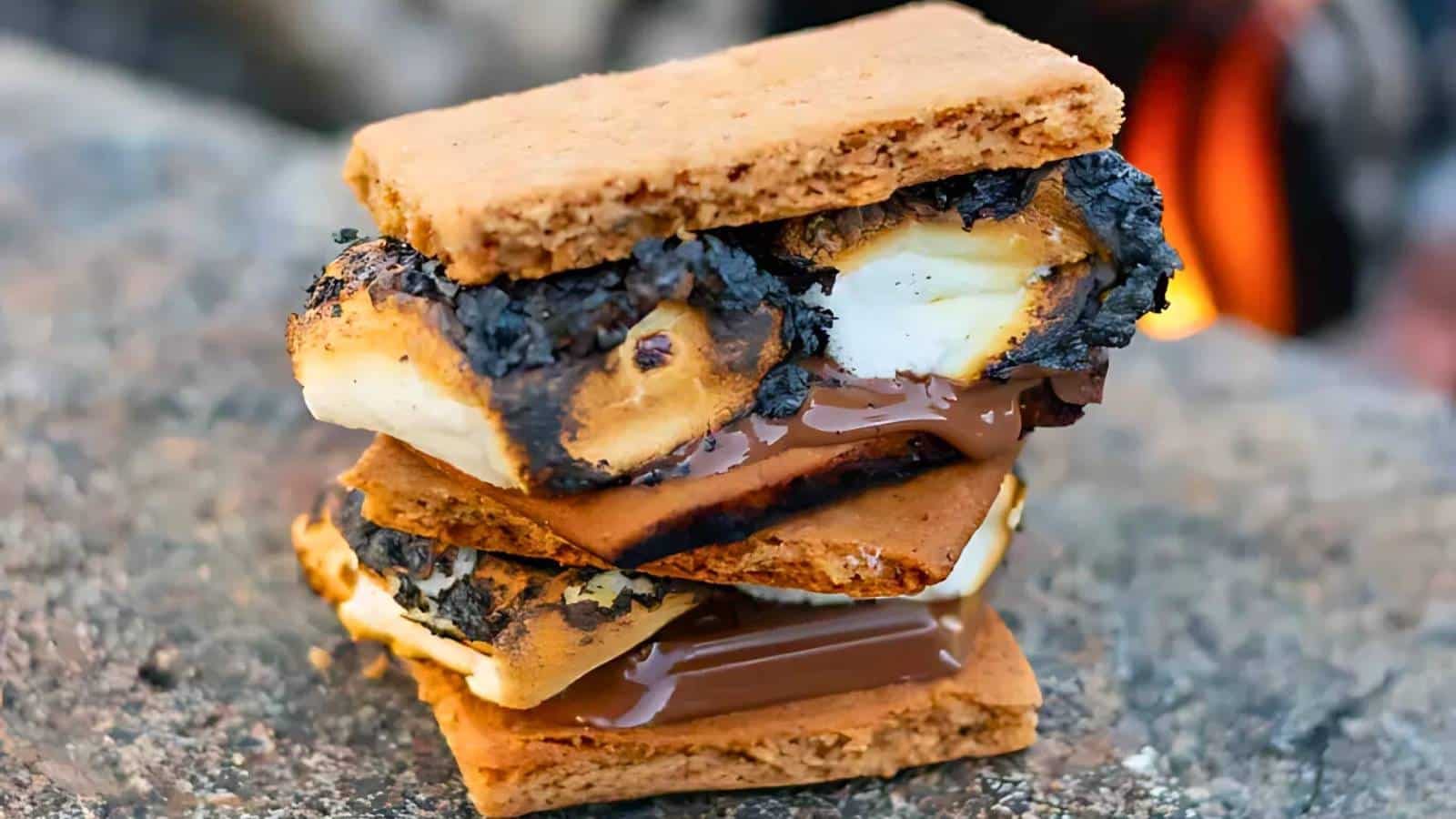 A s'more with charred marshmallows and melted chocolate sandwiched between two graham crackers, resting on a rock.