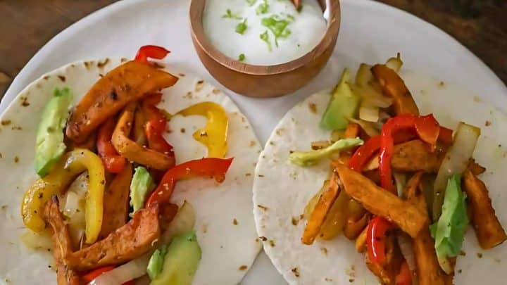 Two tortillas with grilled chicken, red and yellow bell peppers, and avocado slices. A bowl of creamy sauce with green garnish is next to the tortillas on a white plate.