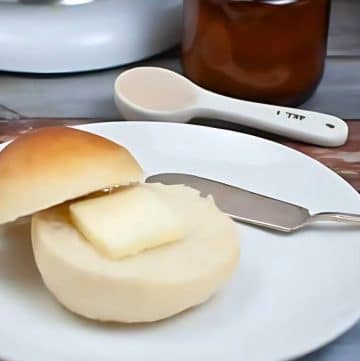 A buttered roll, one of those tasty breads that instantly delights the senses, sits on a plate with a gleaming butter knife. Surrounding it are a polished butter dish, an elegant measuring spoon, and a perfectly placed egg in its holder.
