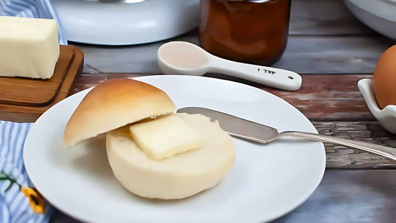 A buttered roll, one of those tasty breads that instantly delights the senses, sits on a plate with a gleaming butter knife. Surrounding it are a polished butter dish, an elegant measuring spoon, and a perfectly placed egg in its holder.