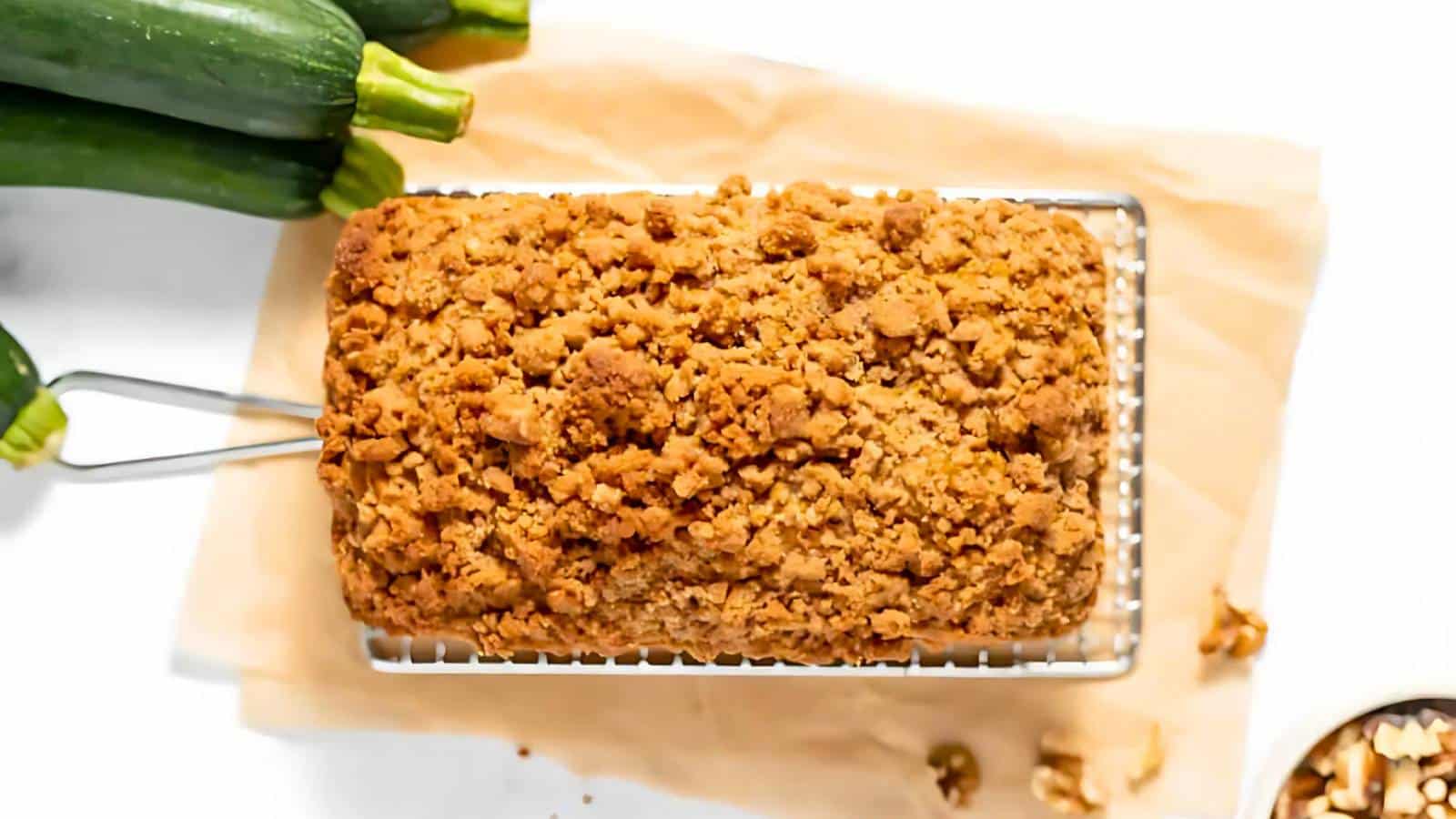 A loaf of zucchini bread with a crumb topping on a cooling rack, surrounded by fresh zucchinis and walnuts on a parchment paper.