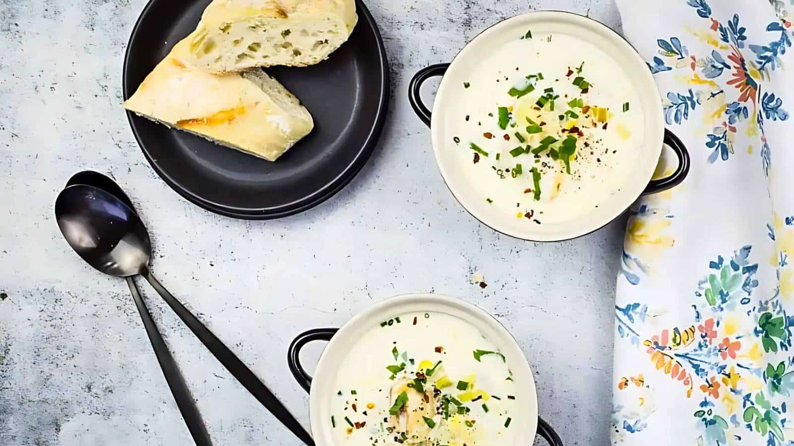 Two bowls of creamy soup garnished with herbs, accompanied by a plate of sliced bread. Two spoons and a floral napkin complete the arrangement.