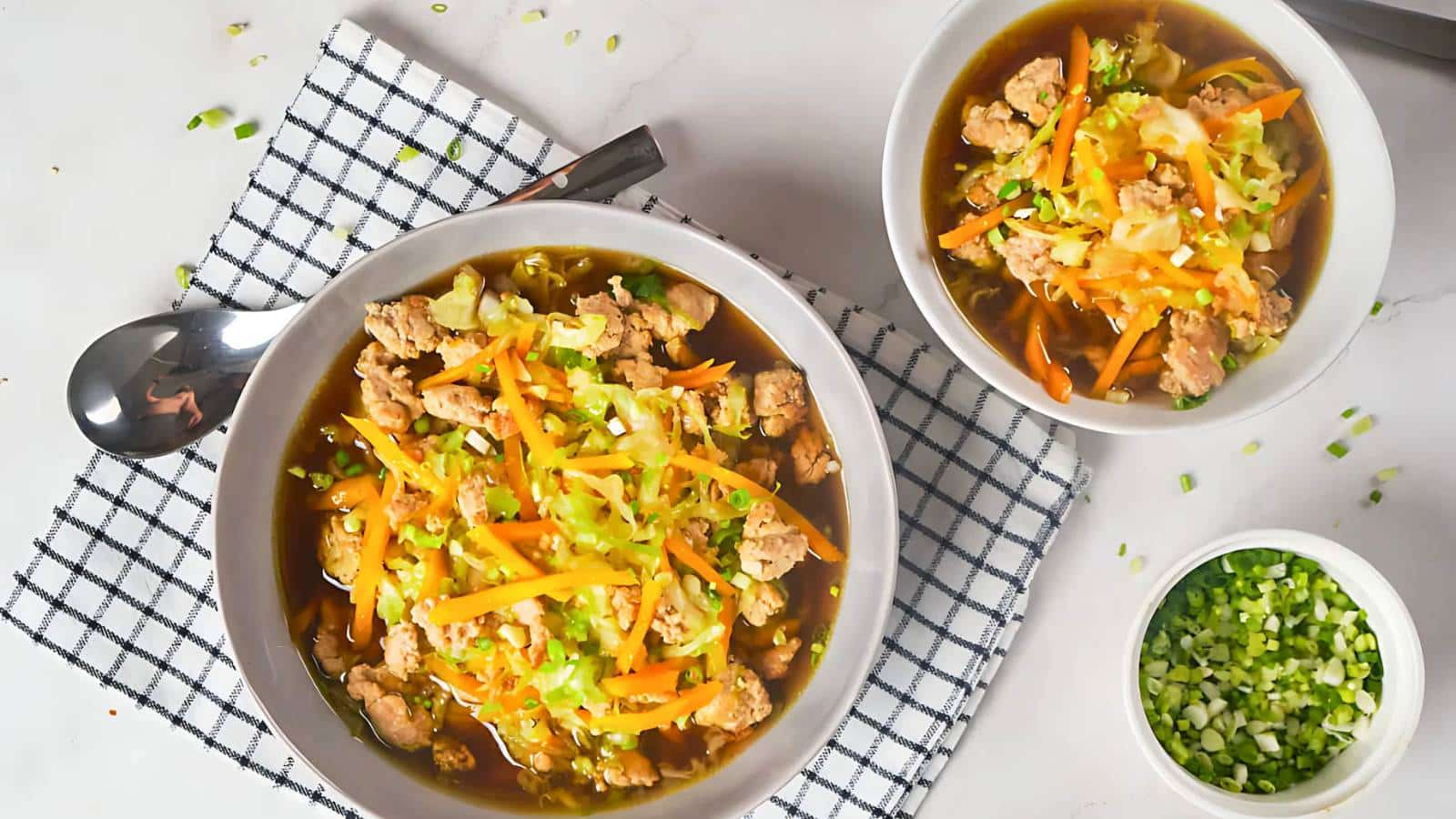 Two bowls of egg roll in a bowl, featuring ground meat, shredded carrots, and cabbage on a checked napkin. A side of chopped green onions is visible.