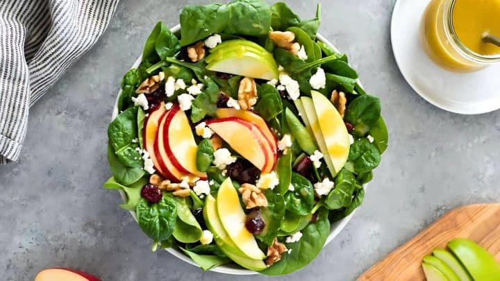 A spinach salad with apple slices, walnuts, cranberries, and feta cheese, next to a jar of dressing.