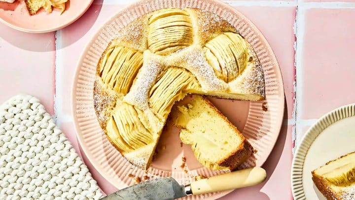 A sliced apple cake on a pink plate, with a knife beside it. The cake is topped with apple slices and dusted with powdered sugar.