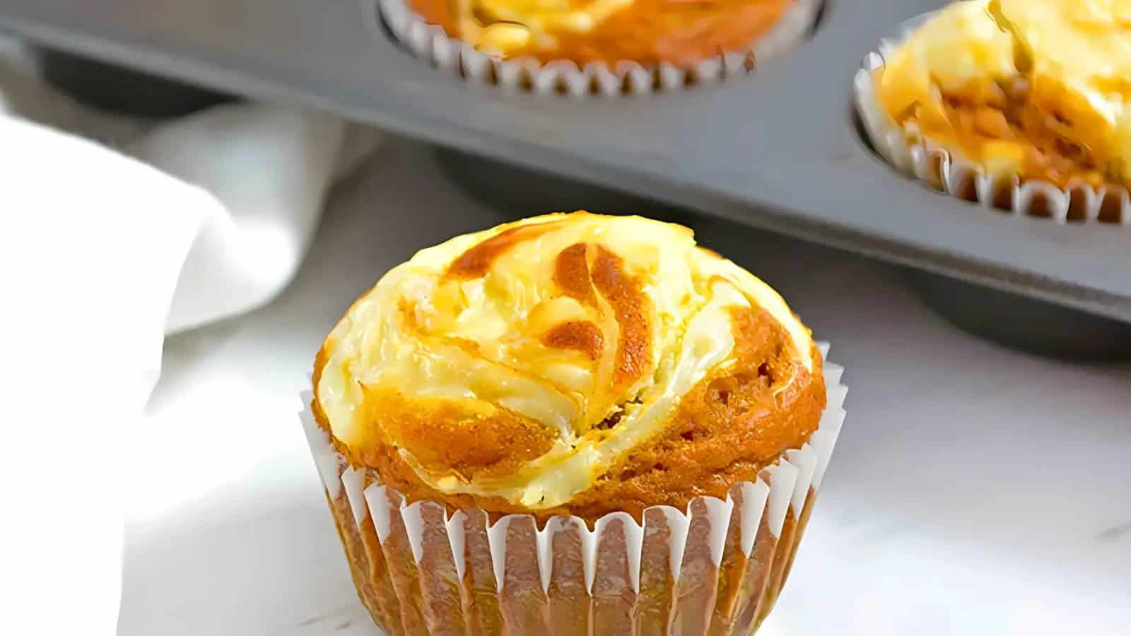 Close-up of a freshly baked muffin in a paper liner with a swirl of cream cheese on top, with more muffins in a baking tray in the background.