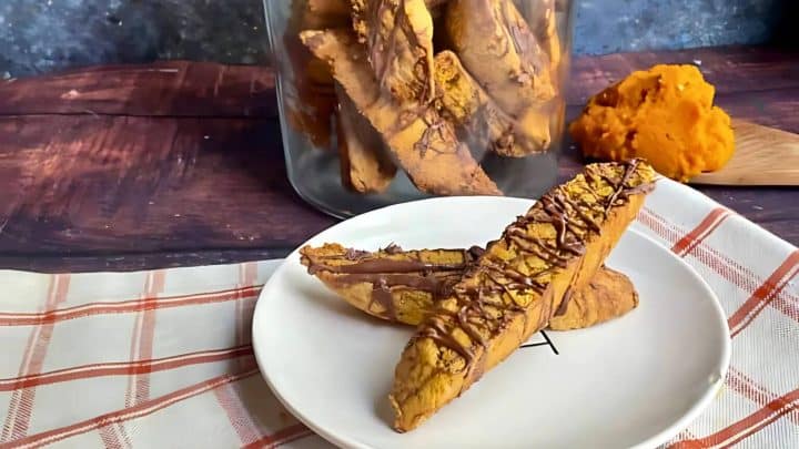 A plate with two chocolate-drizzled biscotti in the foreground. A jar of biscotti and a spoonful of pumpkin puree on a wooden board are in the background.