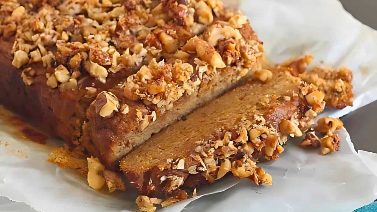 A loaf of bread topped with chopped nuts, partially sliced on parchment paper.