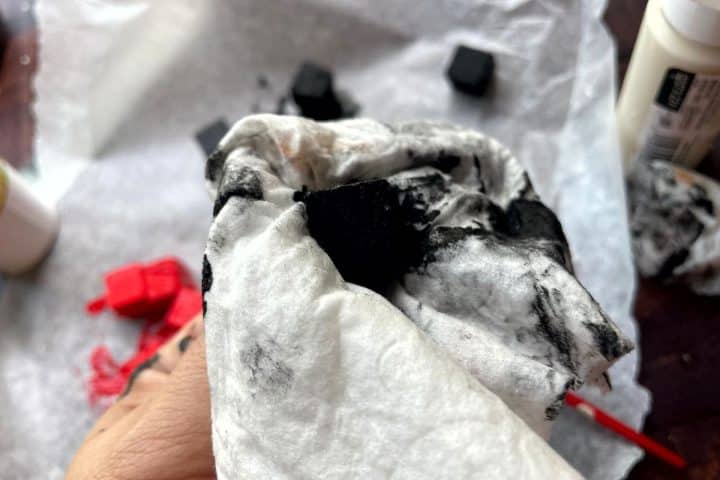 Hand holding a crumpled, black-stained tissue with paint and small black foam cubes on a work surface in the background.