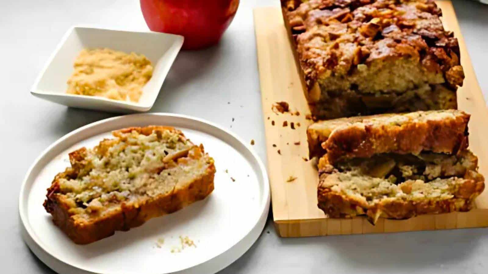 A sliced loaf of apple bread on a cutting board, with a piece on a plate, an apple, and a dish of butter nearby.