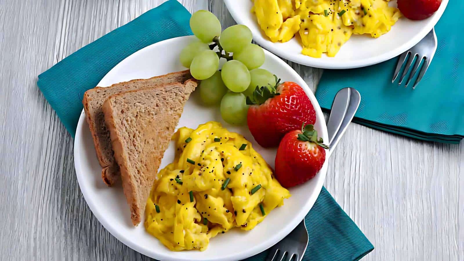 Plates with scrambled eggs, toast, grapes, and strawberries on teal napkins with forks.