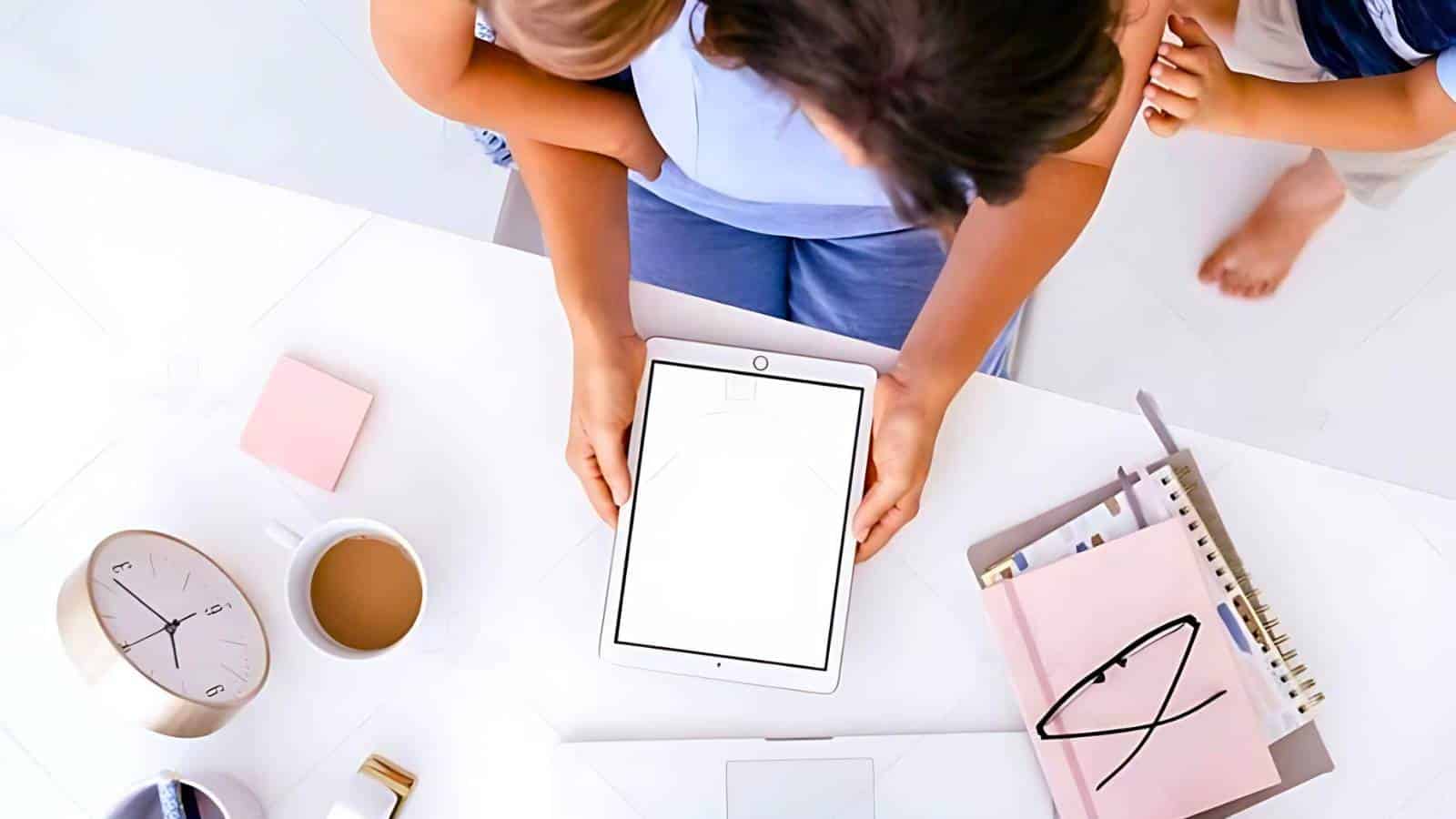 Person using a white tablet at a cluttered desk with a child on their lap. Nearby are a clock, stack of notebooks, coffee mug, and pink sticky notes.