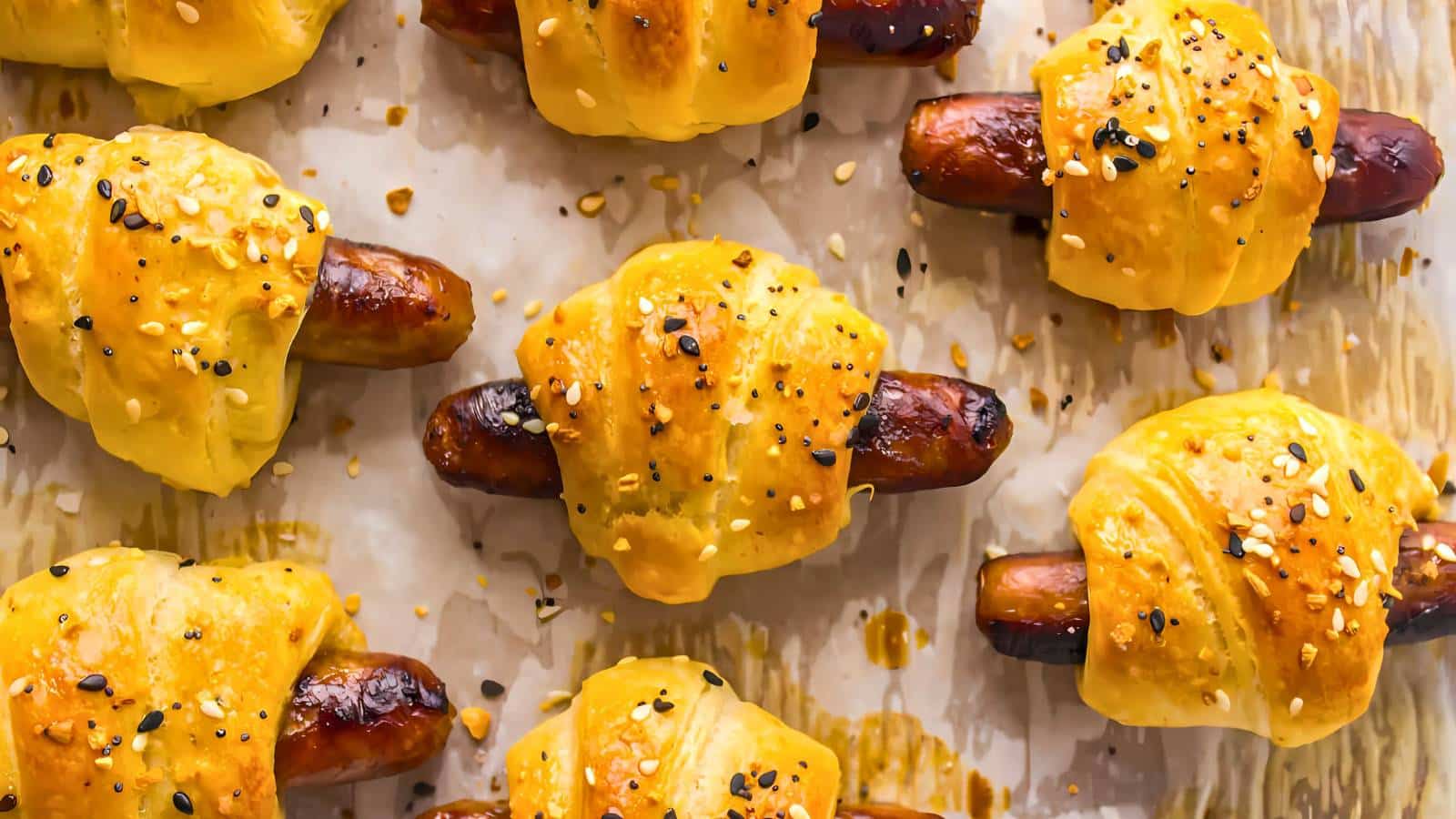 Baked sausages wrapped in golden brown pastry, sprinkled with sesame and poppy seeds, on parchment paper.