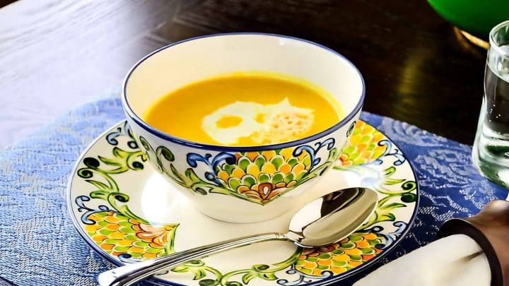 A cup of yellow soup with cream on top, served in a decorative bowl on a matching saucer, with a silver spoon beside it on a blue lace table mat.