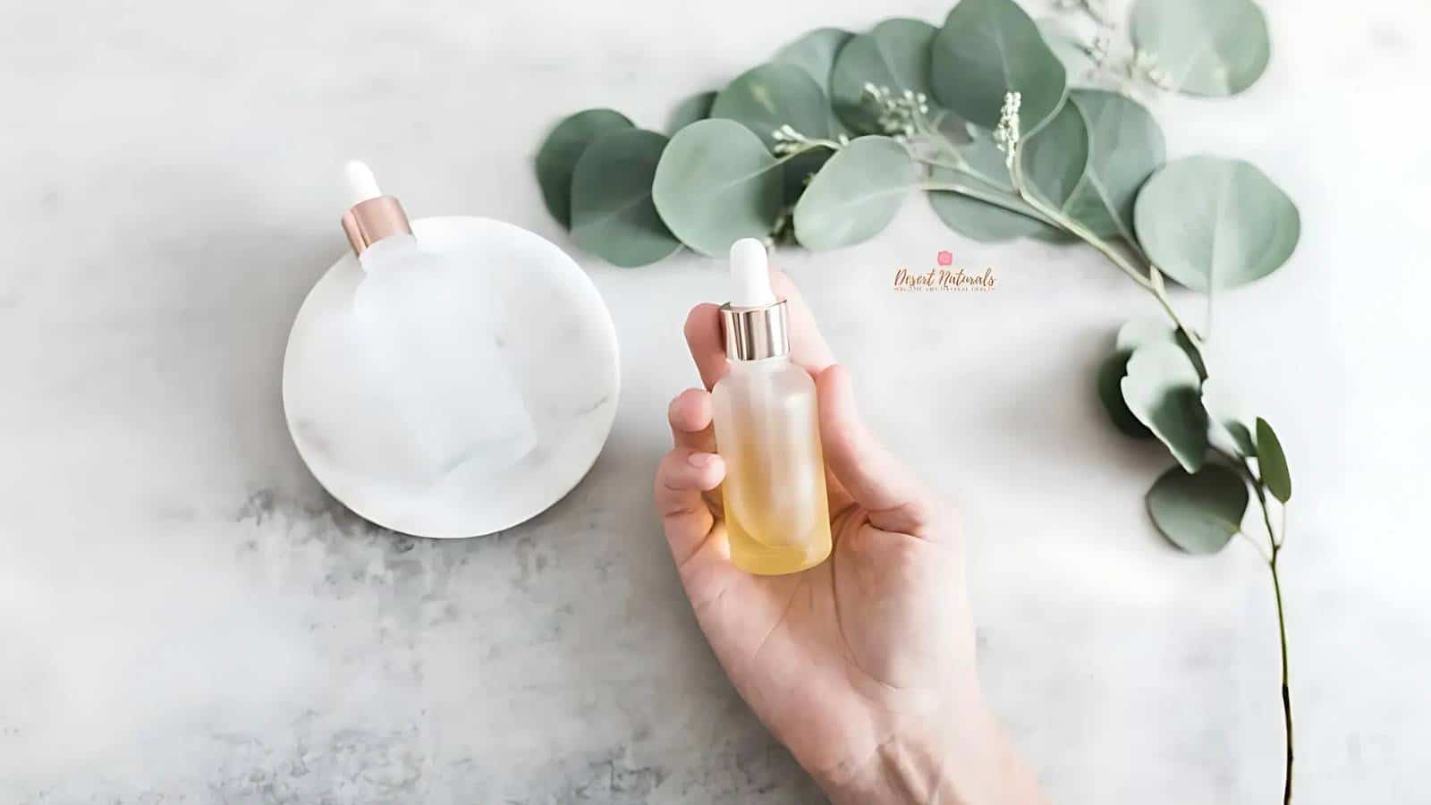 Hand holding a dropper bottle with yellow liquid, next to a frosted bottle on a marble surface. Eucalyptus leaves are placed beside the bottles.