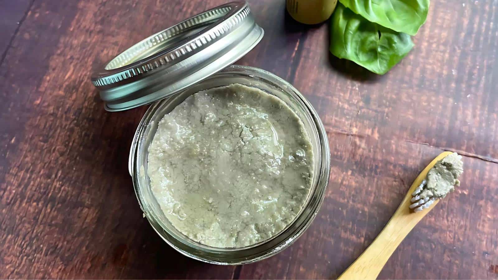 A jar of grayish-green paste with an open metal lid sits on a wooden surface. Nearby are a wooden spoon with paste and some green basil leaves.