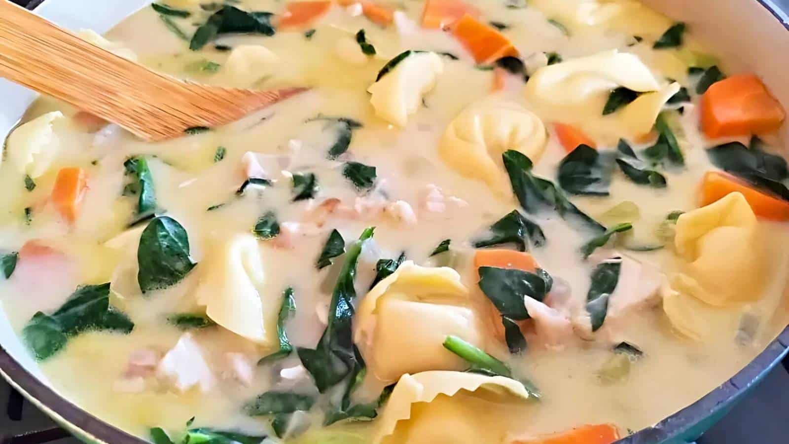 A bowl of creamy soup with vegetables and greens, accompanied by a twisted bread roll on a wooden table.