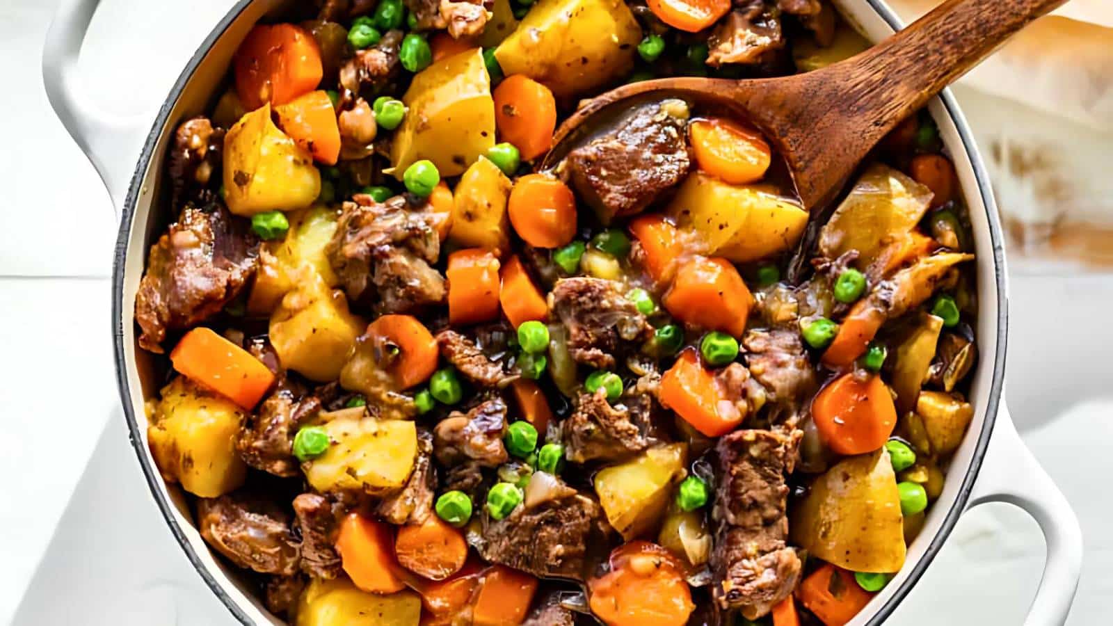 A pot filled with beef stew containing chunks of beef, potatoes, carrots, and peas, with a wooden spoon resting on top.