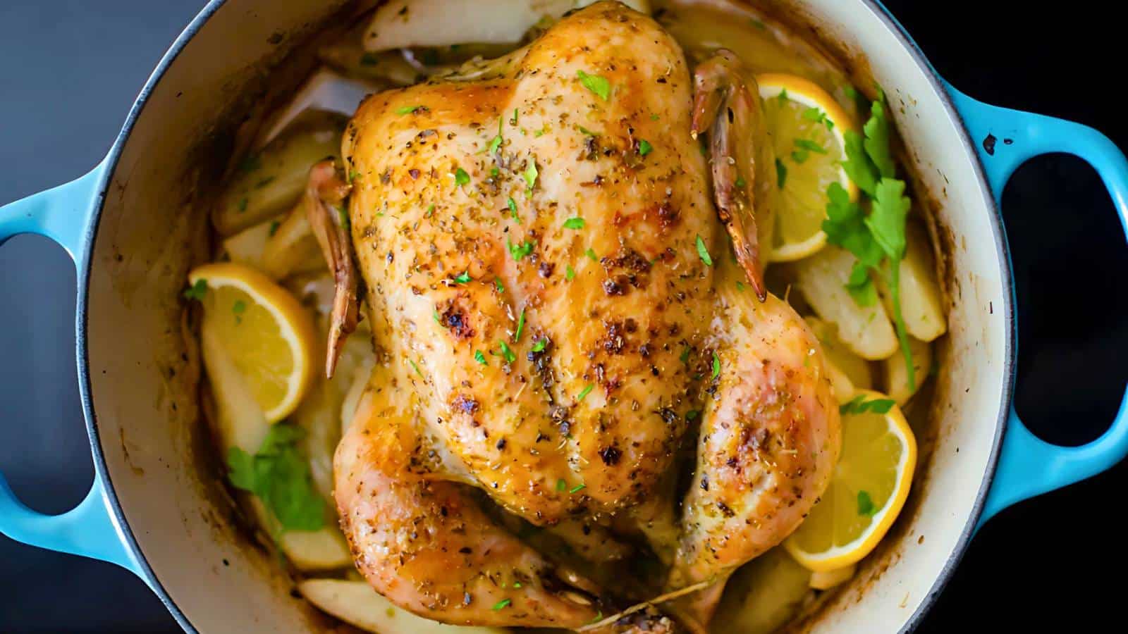 Roast chicken with herbs and lemon slices in a blue pot, surrounded by potato wedges.
