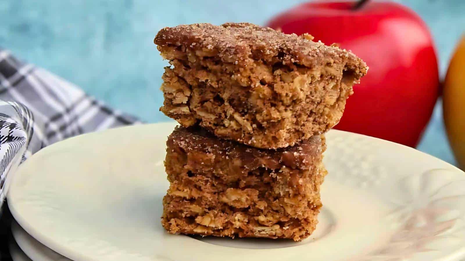 Two stacked oatmeal cake squares on a plate, with a red apple in the background.