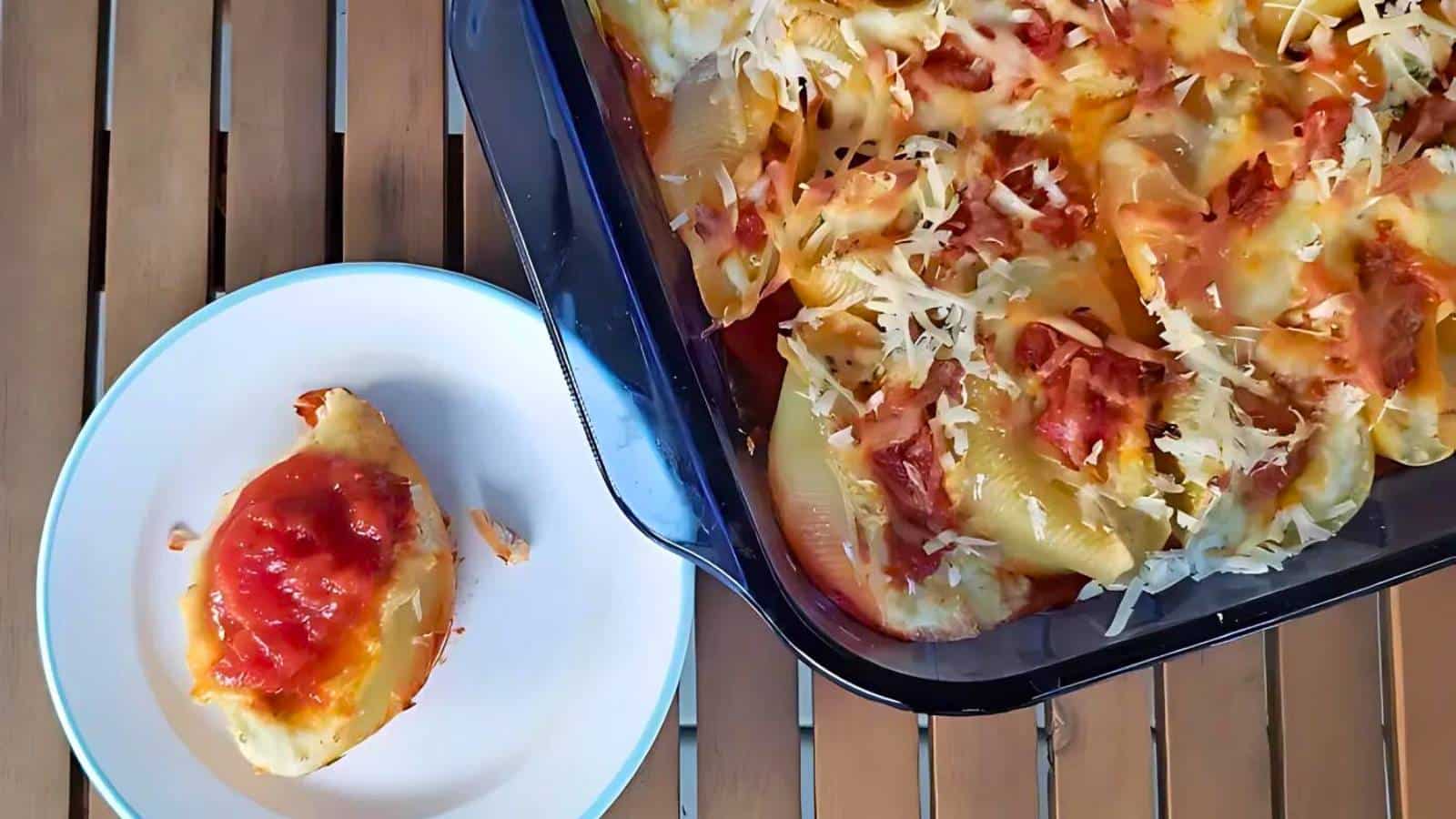 A baking dish filled with cheesy stuffed pasta shells topped with tomato sauce sits on a slatted wood table. A single stuffed shell with sauce is served on a white plate next to it.