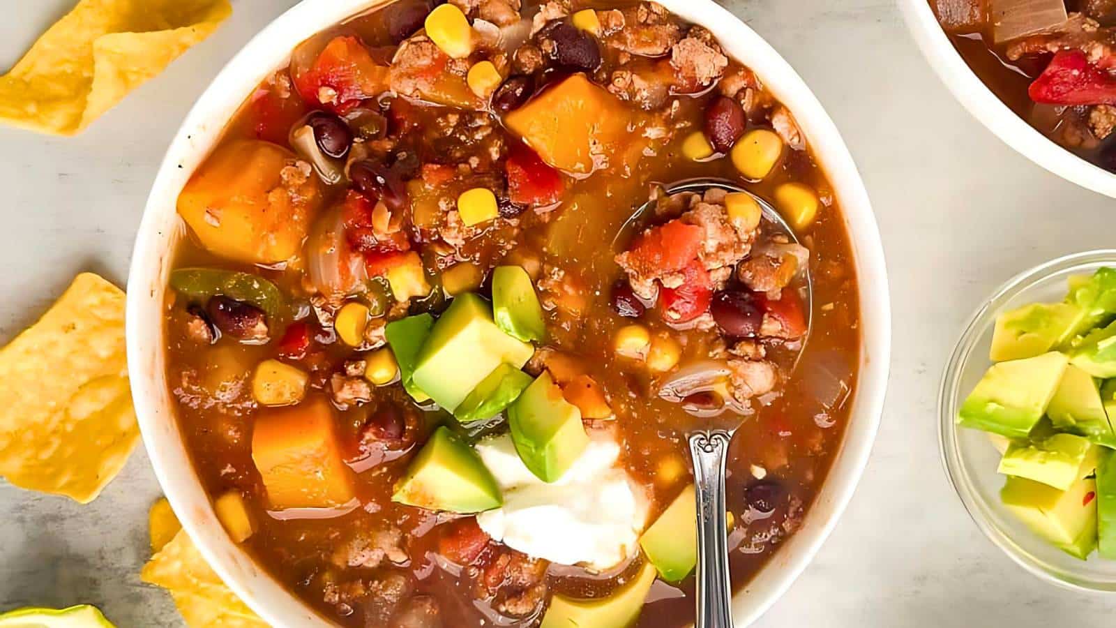 A bowl of hearty soup filled with chunks of sweet potato, black beans, corn, tomatoes, and ground meat, topped with avocado and a dollop of sour cream, with tortilla chips on the side.
