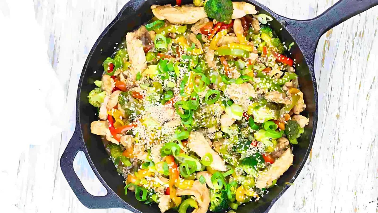 A skillet of stir-fried vegetables with sliced chicken, broccoli, bell peppers, sesame seeds, and green onions.