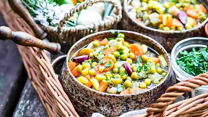 Bowl of mixed vegetable stew with carrots, peas, and herbs, placed in a rustic basket alongside garlic.