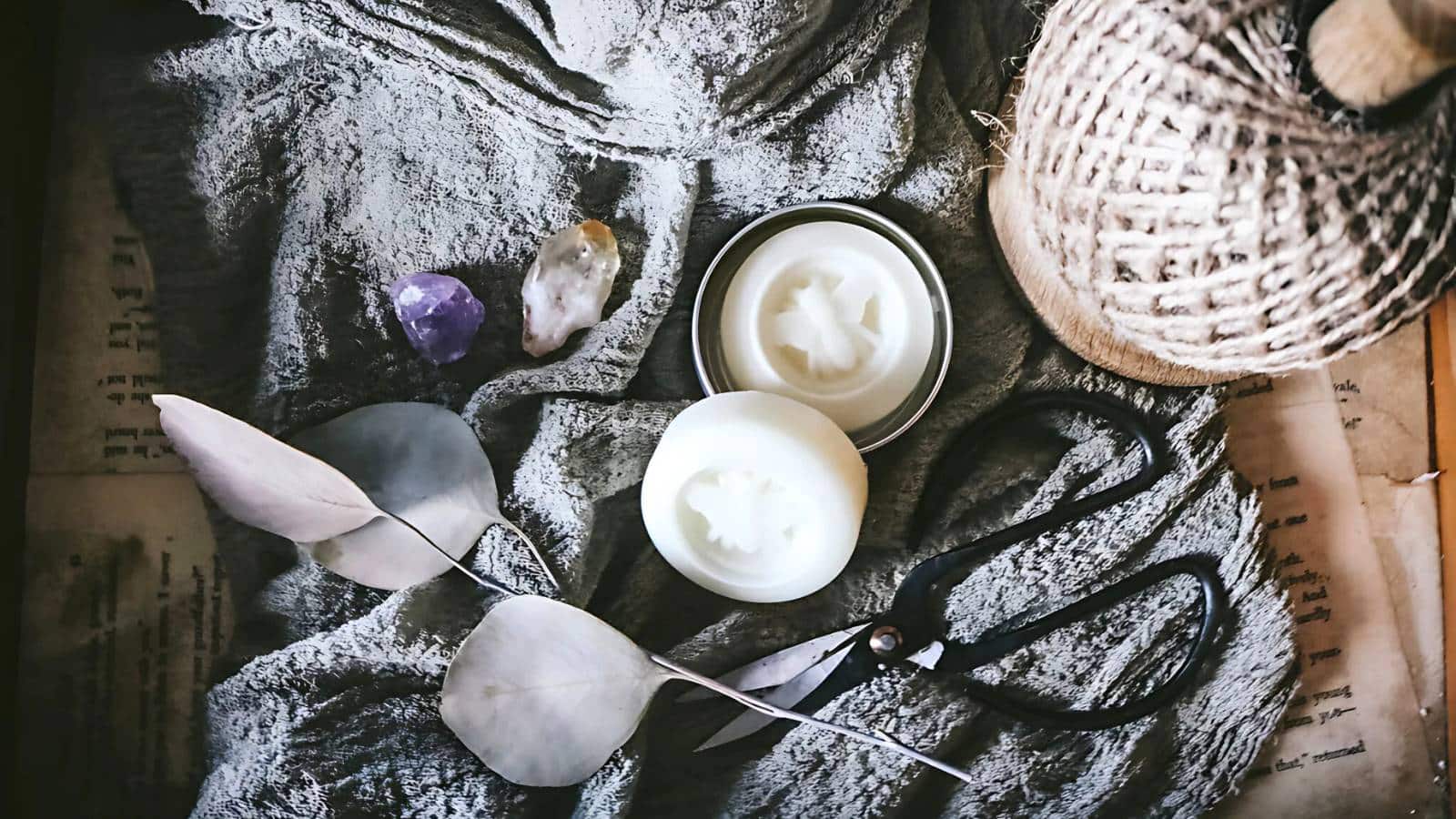 A flat lay of craft supplies: two crystals, a gray cloth, a tin with a white disc, scissors, a ball of twine, dried leaves, and vintage paper.