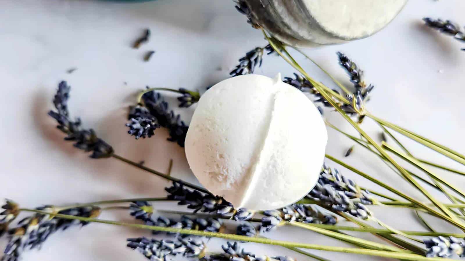 A white bath bomb sits on a surface surrounded by dried lavender sprigs.
