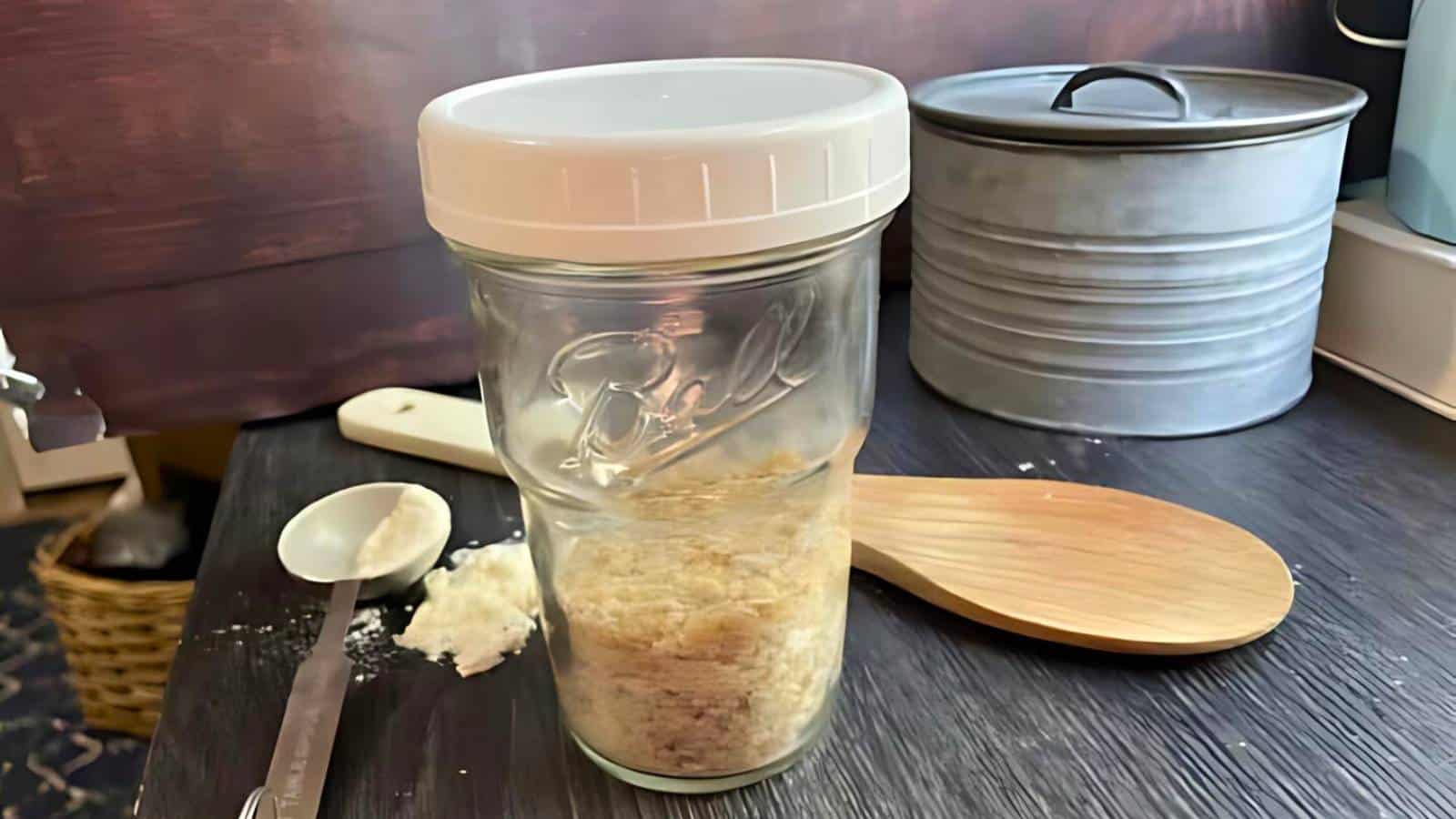 A jar with a white lid partially filled with dry ingredients is on a counter next to a wooden spoon, metal container, and measuring spoon with powder.