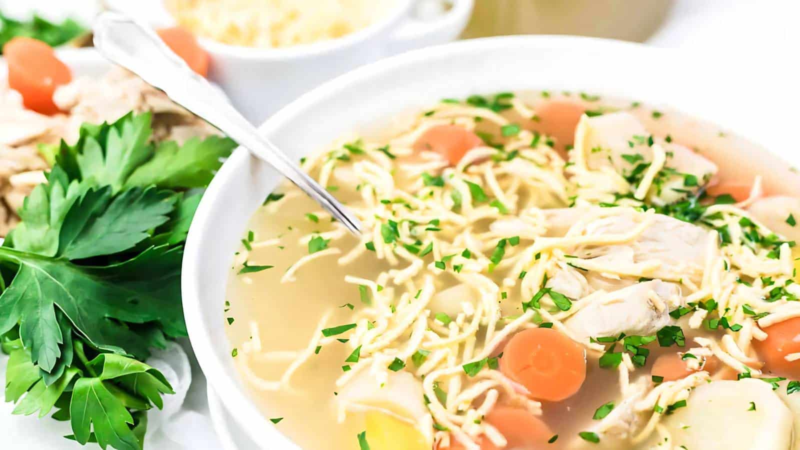 Bowl of chicken noodle soup with carrots and parsley garnish, alongside fresh parsley and a small dish of grated cheese.