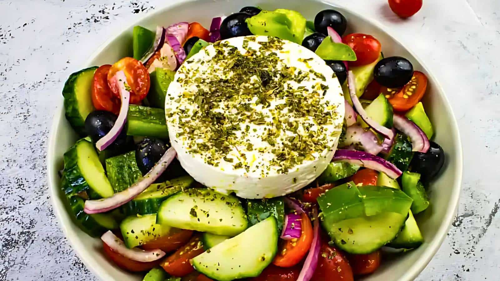 A bowl of Greek salad with sliced cucumbers, tomatoes, red onions, black olives, and a round block of feta cheese topped with herbs.